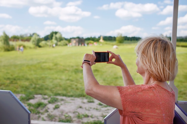 Mujer turista senior feliz tomando fotos en el teléfono inteligente mientras viaja en automóvil turístico en el parque natural