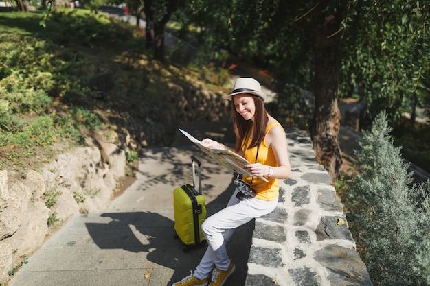 Mujer de turista viajero alegre en ropa casual sombrero con maleta mirando el mapa de la ciudad sentado en piedra en la ciudad al aire libre. Chica viajando al extranjero para viajar en una escapada de fin de semana. Estilo de vida de viaje turístico.