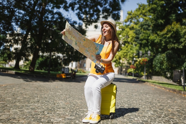 Mujer de turista viajero asombrado en ropa amarilla sombrero sentado en la maleta mirando en la ruta de búsqueda de mapa de la ciudad en la ciudad al aire libre. Chica viajando al extranjero para viajar en una escapada de fin de semana. Estilo de vida de viaje turístico.