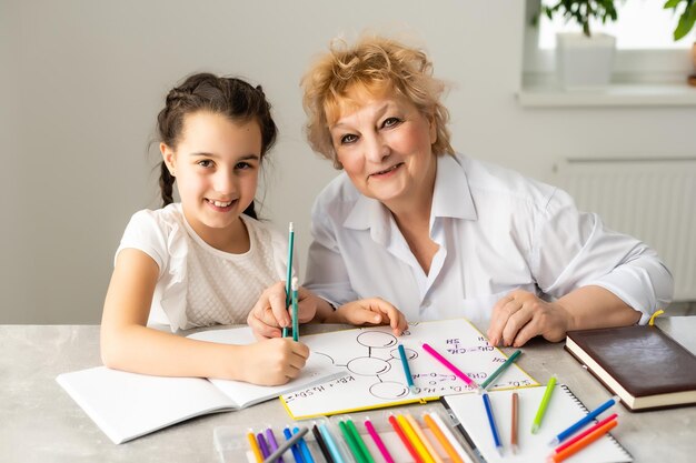 Mujer tutora o madre adoptiva ayudando a una linda niña caucásica a hacer la tarea sentada en la mesa. Diversas niñeras y niños aprendiendo a escribir en un cuaderno estudiando en casa.