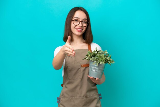 Mujer ucraniana jardinera sosteniendo una planta aislada de fondo azul estrechando la mano para cerrar un buen trato