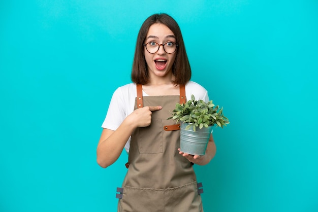 Mujer ucraniana jardinera sosteniendo una planta aislada de fondo azul con expresión facial sorpresa