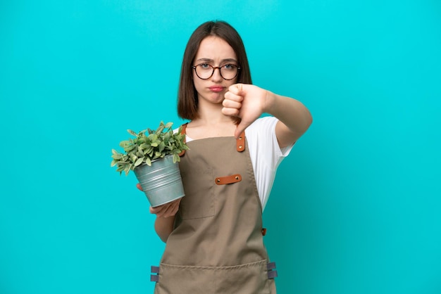 Mujer ucraniana jardinera sosteniendo una planta aislada de fondo azul mostrando el pulgar hacia abajo con expresión negativa