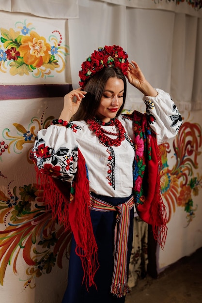 Mujer ucraniana en ropa étnica tradicional y corona floral roja en el fondo de la estufa decorada en la choza Vestido bordado nacional ucraniano llamada vyshyvanka Ore por Ucrania