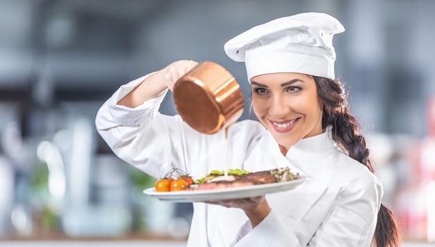 Mujer en uniforme de chef vierte salsa sobre bistec con verduras en la cocina.