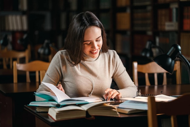 Mujer universitaria sonriente sentada junto a la mesa con una tableta y escribiendo algo en la biblioteca