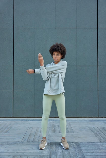 mujer usa chándal estira los brazos ejercicios al aire libre tiene poses de cuerpo atlético contra la pared gris enfocado hacia adelante tiene entrenamiento regular