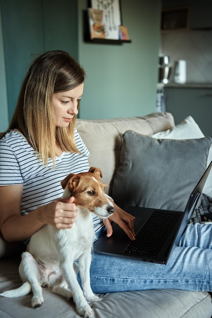 La mujer usa una computadora portátil en la sala de estar y acaricia a su perro trabajando desde casa con una mascota independiente que está sentada