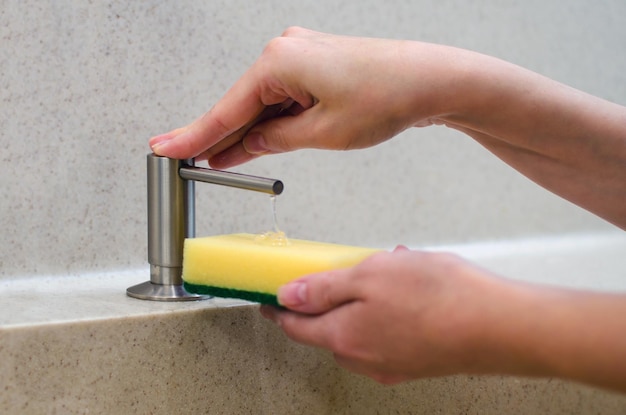 Una mujer usa un dispensador de jabón líquido en la cocina, una esponja para lavar platos. Mezclador de agua cromado brillo. Encimera y fregadero de mármol.