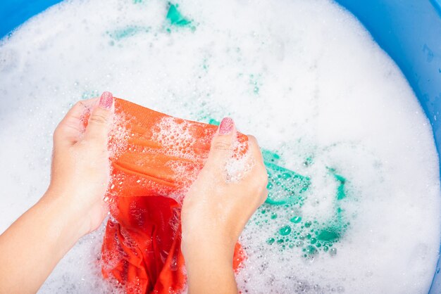 La mujer usa las manos lavando ropa de color en el lavabo con detergente y agua con burbujas de jabón