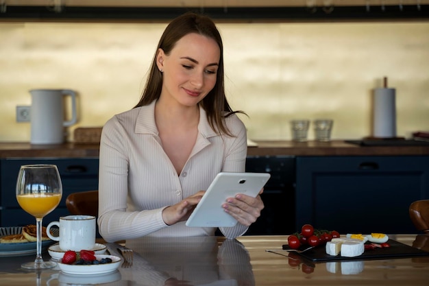 La mujer usa una tableta digital o navega por Internet sentada en una mesa con fruta y queso y una taza