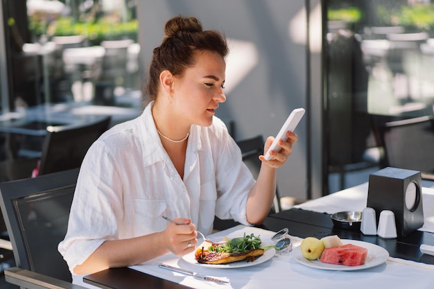 Una mujer usa un teléfono y come el almuerzo o el desayuno al aire libre en una cafetería