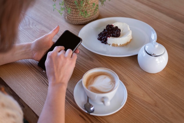 Mujer usa teléfono móvil en café contra mesa capuchino café y postre Smartphone en manos femeninas