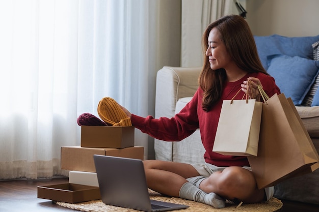 Una mujer usando una computadora portátil para comprar en línea sosteniendo bolsas de compras y abriendo una caja de paquetes postales en casa
