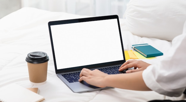 Mujer usando y escribiendo en la computadora portátil con una pantalla blanca de maqueta en la cama.