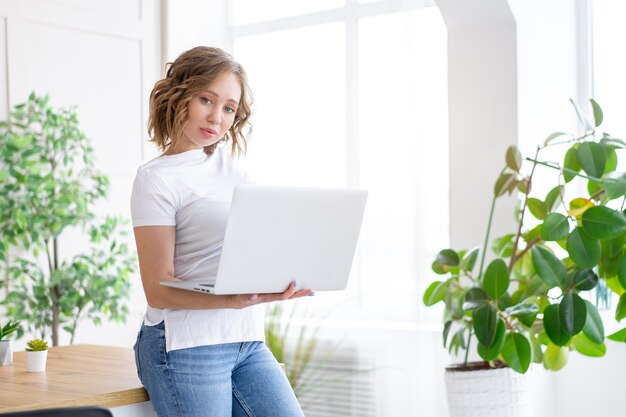 Mujer usando laptop de pie cerca del interior de la oficina blanca de escritorio con planta de interior mirando a cámara