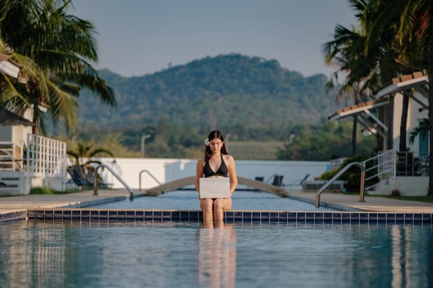 Mujer usando su computadora portátil en el borde de la piscina en un día soleado Trabajo independiente en un país tropical