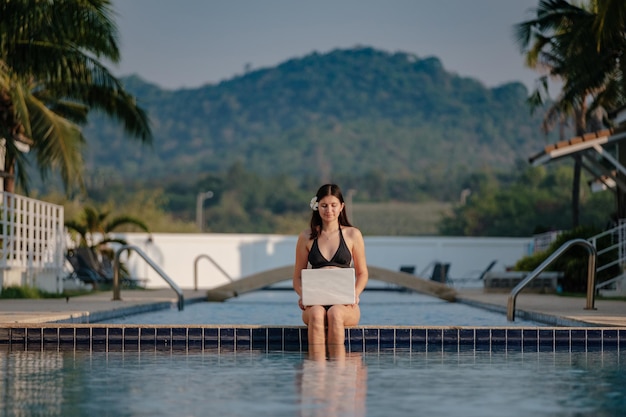 Mujer usando su computadora portátil en el borde de la piscina en un día soleado Trabajo independiente en un país tropical