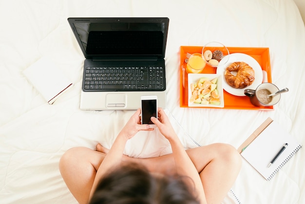 Mujer usando su teléfono móvil y desayunando. Ella esta en el cuarto