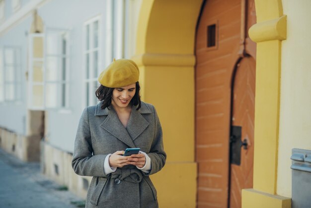 Mujer usando un teléfono inteligente y bebiendo café para llevar mientras usa una boina amarilla