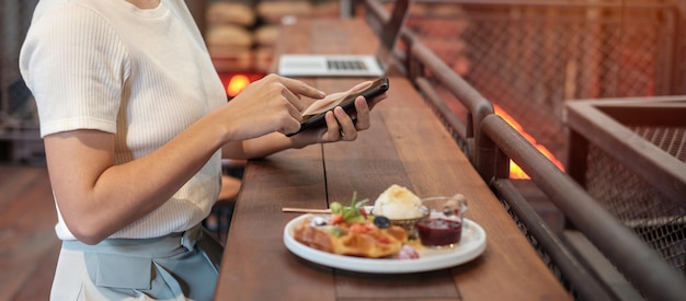 Foto mujer usando teléfono inteligente y comiendo postre con bayas frescas y fruta en el café