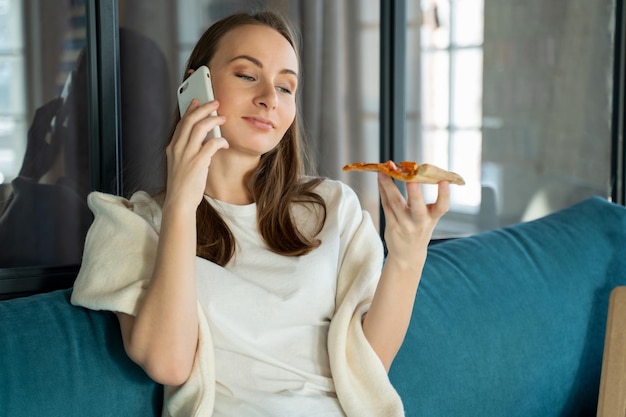 Mujer usando un teléfono inteligente mientras come una rebanada de pizza de una caja mujer joven comiendo pizza en el