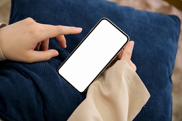 Mujer usando un teléfono inteligente en su sala de estar chateando con la maqueta de pantalla blanca del teléfono de sus amigos