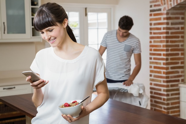 Mujer usando un teléfono móvil y desayunando cereales mientras el hombre plancha una camisa