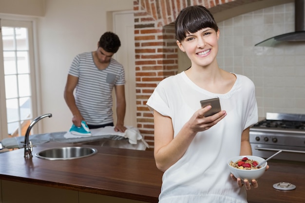 Mujer usando un teléfono móvil y desayunando cereales mientras el hombre plancha una camisa