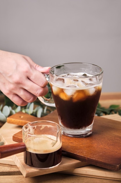 La mujer usó la mano para tomar café frío helado negro en un vaso con trozos de hielo en una mesa de madera, luz natural, bebida fresca de verano por la mañana