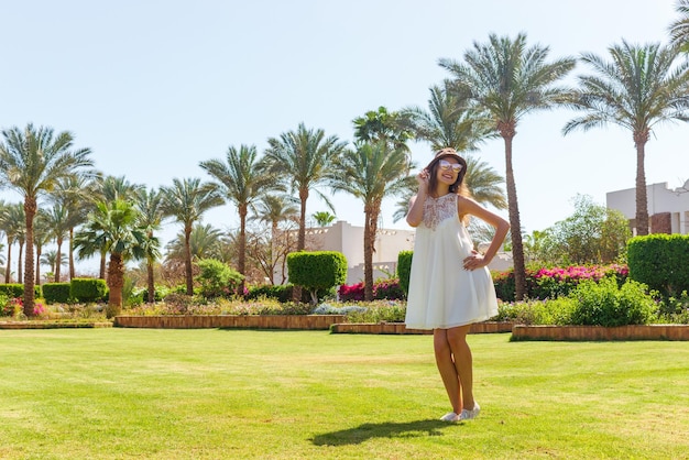 Mujer de vacaciones con gafas de sol y vestido blanco relajándose al aire libre en un hermoso parque verde con palmeras