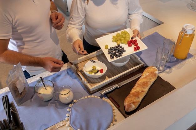 Mujer vaciando frutas en un recipiente con yogur mientras prepara el desayuno con su pareja