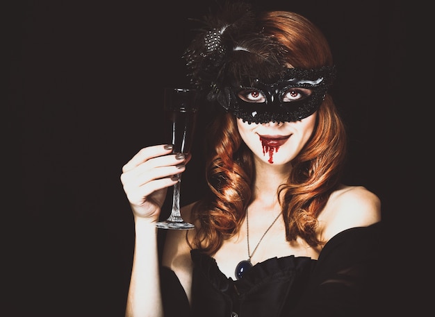 Mujer vampiro pelirroja en máscara con vaso de sangre. Foto de estilo vintage.