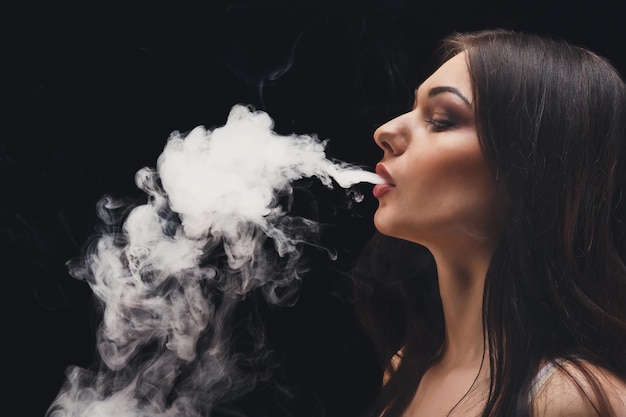 Mujer vaping cigarrillo electrónico con humo sobre fondo negro de cerca. Chica morena que sopla una nube de humo sobre fondo negro. Concepto de vapor y fumar sin nicotina, espacio de copia