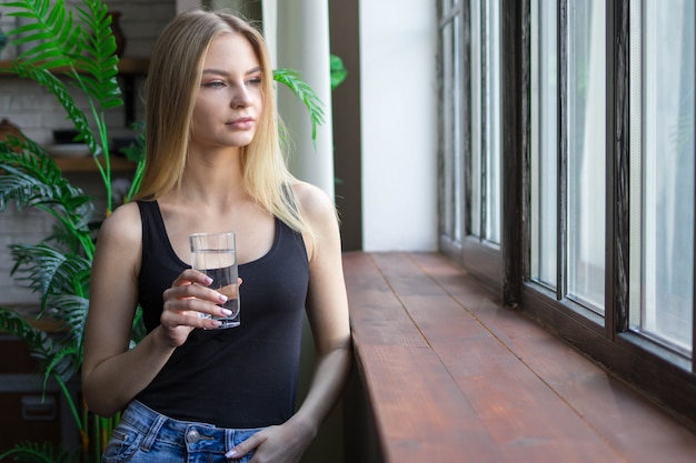 Una mujer con un vaso de agua mira pensativamente por la ventana.