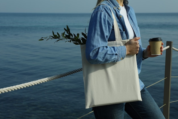 Mujer con vaso de papel y bolsa ecológica de pie en el muelle contra el mar