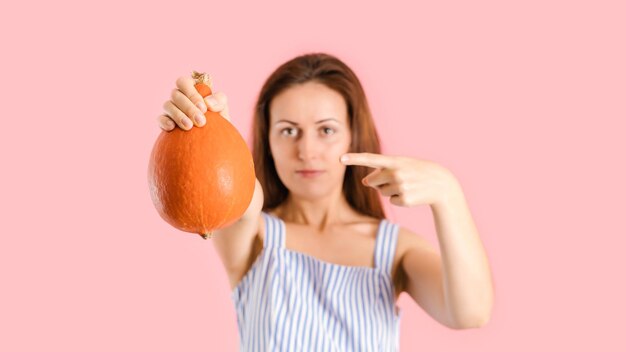 Una mujer vegana sostiene una calabaza en sus manos y la señala con su dedo índice. Foto de estudio sobre un fondo rosa