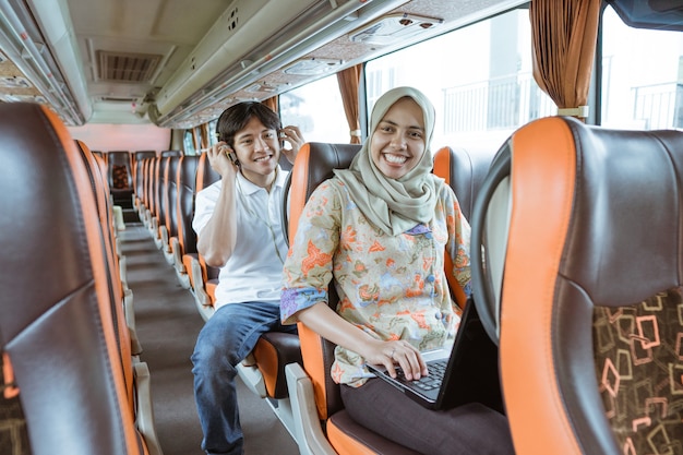 Una mujer con velo sonríe mientras usa una computadora portátil y un joven se sienta detrás de ella mientras usa audífonos en el autobús