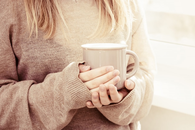 Mujer se para en la ventana y sostiene una taza de bebida caliente