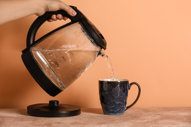 Mujer vertiendo agua hervida caliente de hervidor eléctrico en taza en la mesa