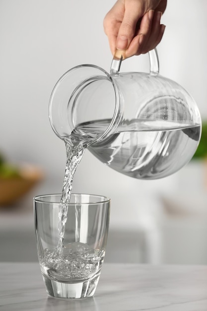 Mujer vertiendo agua de una jarra en un vaso sobre una mesa de mármol blanco en el primer plano de la cocina