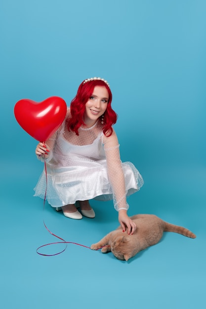 Mujer vestida de blanco sosteniendo un globo volador en forma de corazón, jugando con un gato rojo