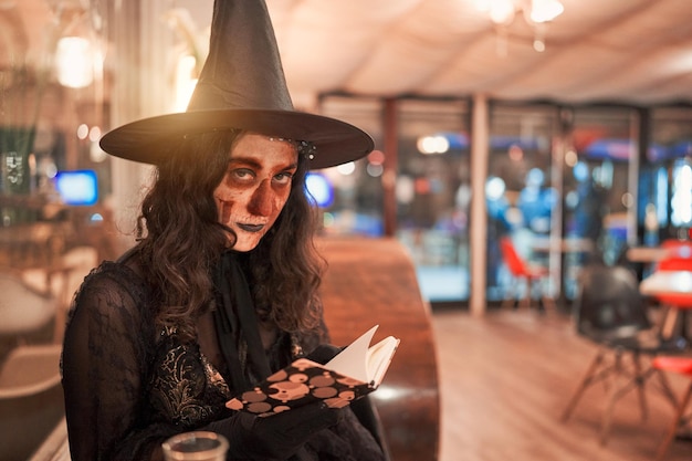 Mujer vestida de bruja y con la cara pintada como una calavera sentada en un café leyendo un retrato de libro