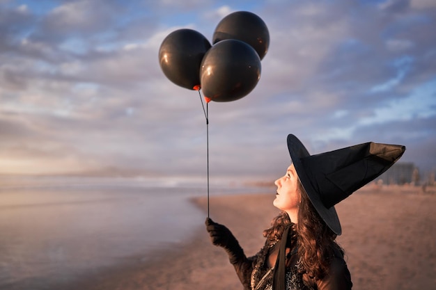 Mujer vestida de bruja sosteniendo globos negros en la playa de perfil