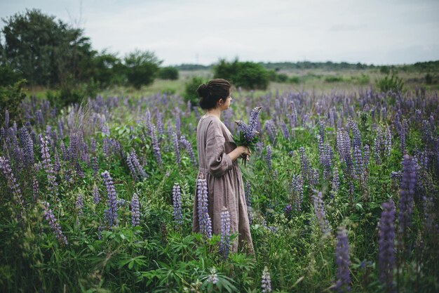 Mujer vestida de forma rústica reuniendo un ramo de lupino en una pradera estética Cottagecore atmosférica