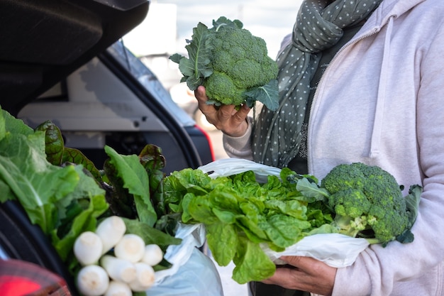 Mujer vestida informal después de comprar verduras frescas colocándolas en el maletero del coche. Sosteniendo un brócoli crudo. Concepto de estilo de vida saludable y vegetariano