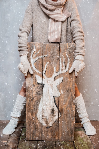 Mujer vestida para el invierno con artículos de moda de punto blanco sostiene un cartel de madera con fondo pintado de ciervo blanco gris.