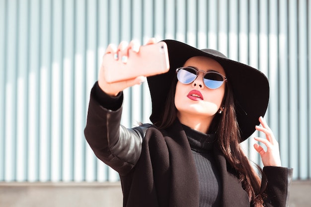 Mujer vestida a la moda lleva sombrero y gafas de sol, tomando selfie en teléfono móvil