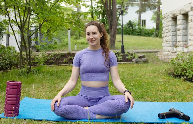 Una mujer vestida de púrpura está sentada en una alfombra de yoga en un parque.