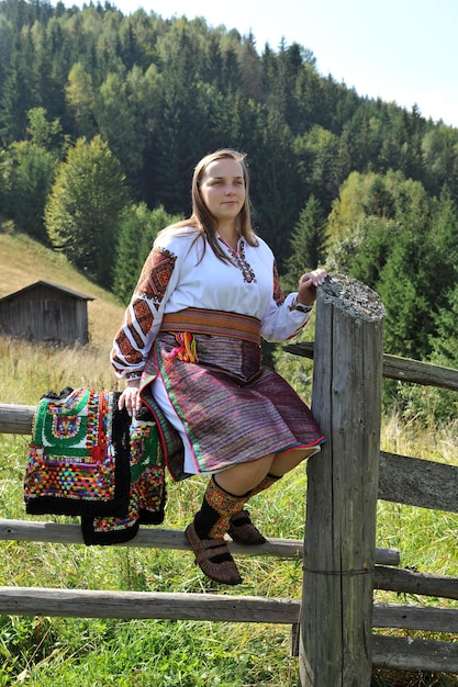 Una mujer vestida con ropa nacional Hutsul se sienta en una valla contra el fondo de las montañas